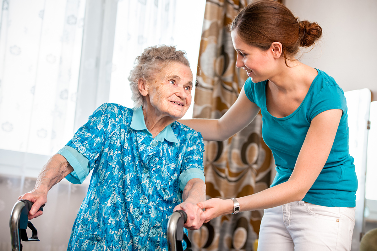 Eine Pflegefachperson legt ihre Hände auf die Gehhilfe einer Seniorin und sie schauen sich dabei an.